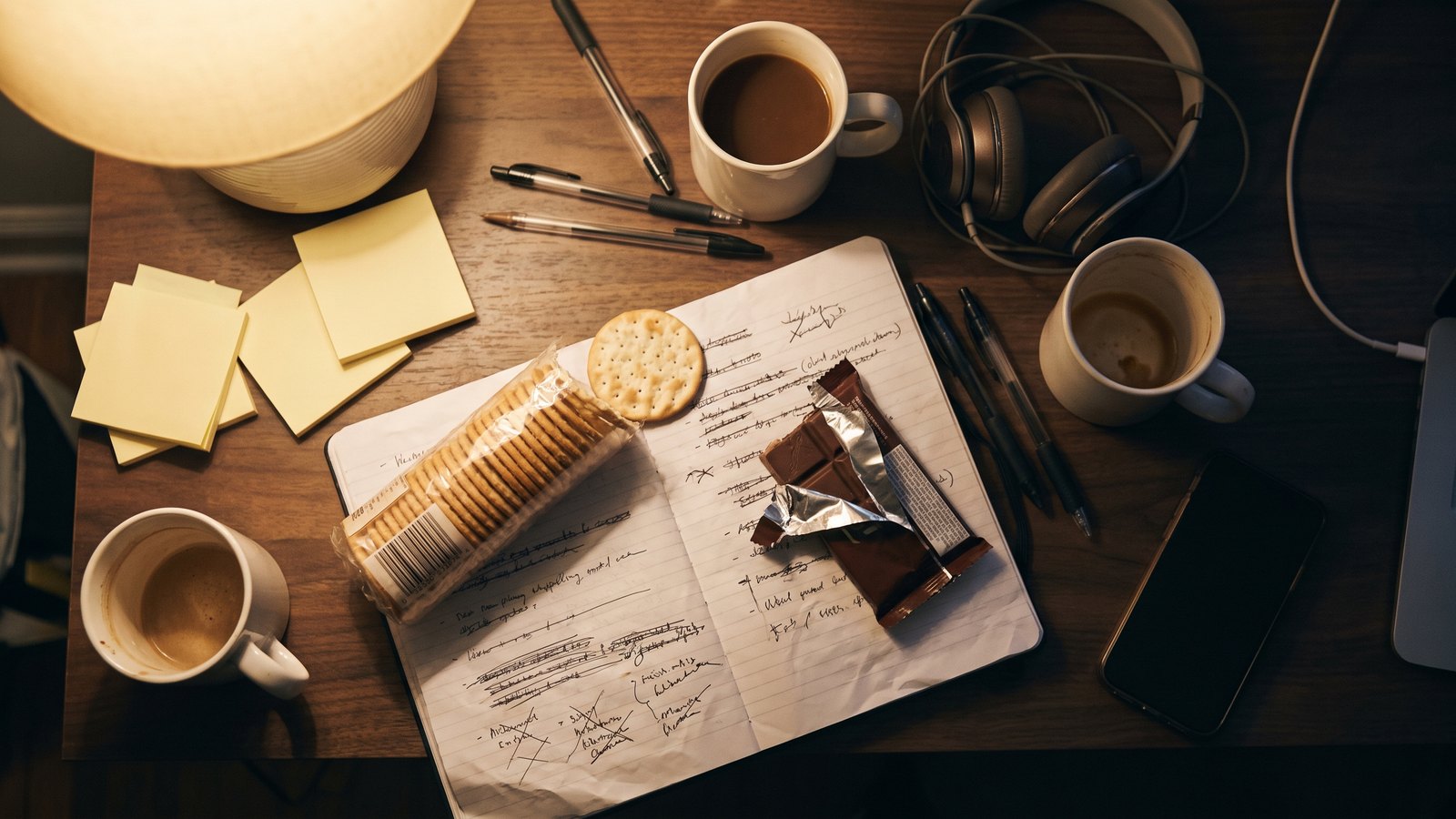 Overhead view of a cluttered desk at dusk with a crossed-out to-do list, an open sleeve of crackers, a part-eaten chocolate bar and three unfinished mugs of coffee.