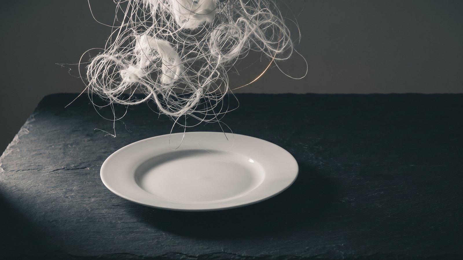 A plain white dinner plate on a dark slate table with a dense tangle of pale thread swirling in the air above it, lit from one side.
