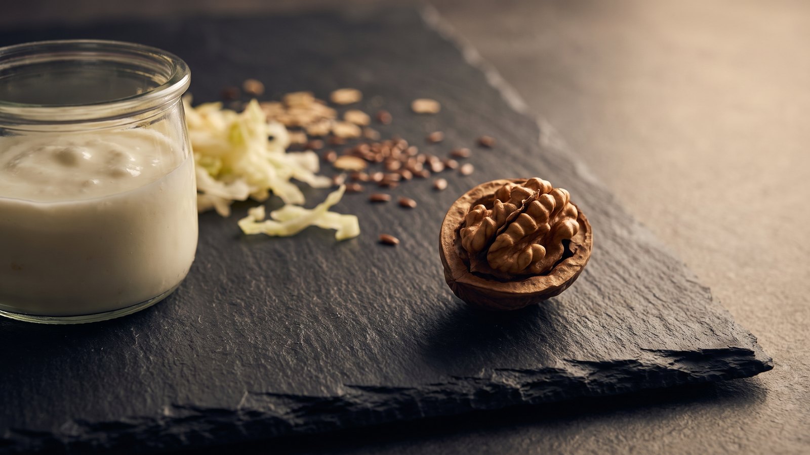 Editorial still life on dark charcoal slate of a walnut half showing its brain-like interior, beside a glass jar of plain yogurt, shredded fermented cabbage, oats and flax seeds